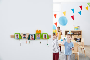 Fünf Kinder auf einer Geburtstagsfeier in einem Klassenzimmer, einige mit Partyhüten und Kronen, stehen an einem Tisch und pusten Seifenblasen. An der geschmückten Wand hängen Kinderfotos in bunten Rahmen, und darüber hängen bunte Wimpel.