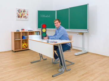 Ein Mann sitzt an einem Schreibtisch in einem Klassenzimmer mit einer grünen Kreidetafel, einem Whiteboard, Regalen mit Spielzeug und einer Ampel mit roten, gelben und grünen Gesichtern. Er lächelt und schaut in die Kamera.