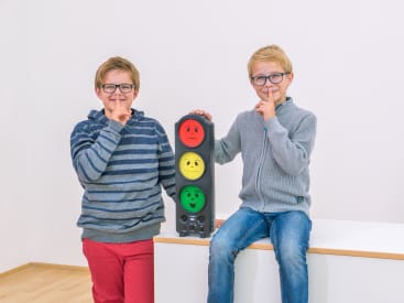 Zwei Jungen mit Brillen halten ein Gerät in Form einer Ampel mit roten, gelben und grünen Gesichtern. Beide Jungen halten einen Finger an die Lippen, um Ruhe zu signalisieren. Sie befinden sich in einem Innenraum mit einem schlichten weißen Hintergrund.