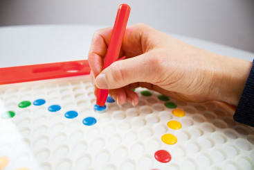 Eine Hand setzt einen blauen runden Stift auf eine weiße Tafel mit gleichmäßig verteilten Löchern. Andere bunte Stifte, darunter gelbe, grüne und rote, sind bereits auf dem Brett positioniert.