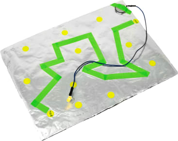 Ein Blatt Alufolie mit einem grünen Zickzackpfad aus Klebeband, mehrere gelbe kreisförmige Aufkleber und eine kleine LED-Leuchte, die über Drähte mit einer Batterie verbunden ist.