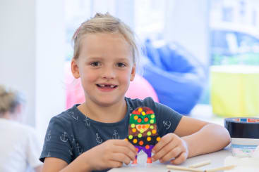 Ein junges Mädchen mit blondem Haar lächelt und hält ein buntes Bastelmonster aus Papier hoch. Sie sitzt an einem Tisch mit Bastelmaterial, und der Hintergrund ist hell und unscharf.