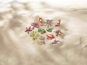 Plastikspielzeug-Meerestiere, darunter ein Seepferdchen, ein Oktopus, ein Delfin, ein Seestern, eine Schildkröte, eine Krabbe und andere, sind in einer Gruppe auf glattem, sonnenbeschienenem Sand mit sanften Schatten angeordnet.