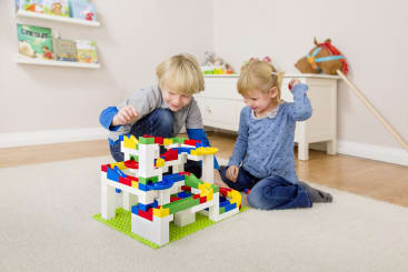 Zwei kleine Kinder sitzen auf einem Teppichboden und spielen mit einer bunten Murmelbahn aus ineinander greifenden Plastiksteinen in einem hellen, aufgeräumten Zimmer mit Regalen und Spielzeug im Hintergrund.