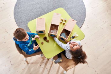 Zwei kleine Kinder sitzen an einem grünen Tisch und spielen mit Holzkisten, die mit verschiedenen natürlichen Gegenständen wie Tannenzapfen und Steinen gefüllt sind. Die Szene ist von oben auf einem hellen Holzboden mit einem grauen Teppich in der Nähe zu sehen.