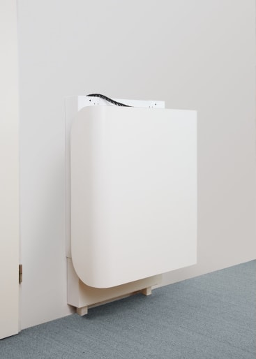 Ein weißer, klappbarer, an der Wand montierter Tisch oder Schreibtisch wird in geschlossener Position vor einer hellen Wand und einem Teppichboden gezeigt. Oben ist ein kleiner Teil eines schwarzen Kabels zu sehen.