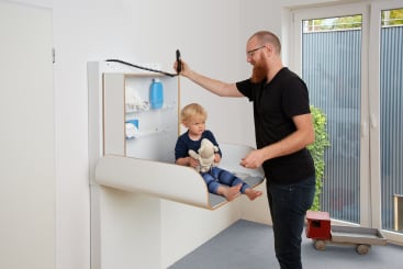Ein Mann steht neben einem Kleinkind, das auf einem an der Wand befestigten Wickeltisch sitzt. Das Kind hält ein Stofftier in der Hand, und in den Regalen darüber liegen Windeln und Zubehör. Der Mann hält einen schnurgebundenen Telefonhörer in der Hand. Auf dem Boden neben einer Glastür steht ein Spielzeugauto.