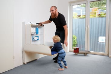 Ein Mann hilft einem Kleinkind in einem Raum mit einer Glastür und einem grauen Teppich, sich neben einem an der Wand montierten Klappwickeltisch aufzustellen. Auf dem Tisch liegen Tücher, Windeln und eine Flasche. Auf dem Boden steht ein kleiner Spielzeuglastwagen.