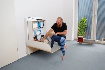 Ein Erwachsener und ein Kleinkind interagieren mit einem herunterklappbaren Wickeltisch, der an einer Wand befestigt ist. Auf dem Tisch befinden sich Babyartikel und Spielzeug. Der Raum hat einen blauen Teppich, eine Topfpflanze, einen Spielzeugwagen und ein großes Fenster.