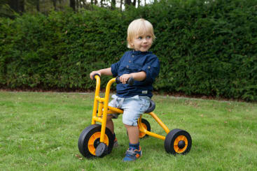 Ein kleines Kind mit blondem Haar sitzt auf einem gelben Dreirad auf einem grasbewachsenen Rasen und trägt ein blaues Hemd, hellblaue Shorts und blaue Turnschuhe. Im Hintergrund sind eine grüne Hecke und Bäume zu sehen.