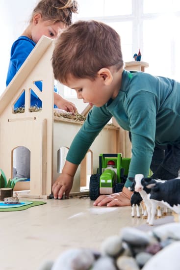 Ein kleiner Junge spielt mit Spielzeugkühen und einem grünen Spielzeugtraktor neben einer hölzernen Spielzeugscheune, während ein Mädchen in einem blauen Hemd mit Stroh in der Scheune spielt. Steine und weitere Spielzeugtiere sind auf dem Boden zu sehen.