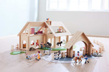 Ein Spielzeugbauernhof aus Holz mit Gebäuden, Zäunen, Pferden, Kühen, Schafen und Figuren von Menschen und Tieren, die auf einem hellen Holzboden in einem hellen Raum mit großen Fenstern angeordnet sind.