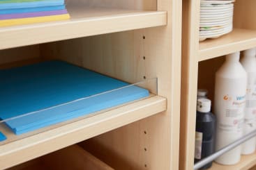 Ein Regal enthält einen Stapel blaues Papier hinter einer durchsichtigen Trennwand. Darüber liegen farbige Papierbögen, und rechts davon befinden sich in den unteren Regalen Teller und Flaschen mit Farbe oder ähnlichen Flüssigkeiten.