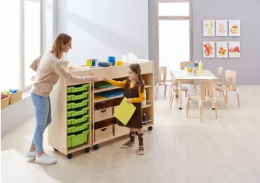 Ein Erwachsener und ein junges Mädchen stehen in einem hellen Klassenzimmer vor einem Regal mit grünen Fächern. Das Mädchen hält ein gelbes Blatt Papier in der Hand. Im Hintergrund sind Kunstzubehör, Tische, Stühle und Kunstwerke der Kinder zu sehen.