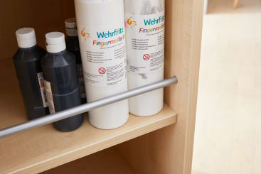 Mehrere Flaschen mit Farbe, darunter weiße und schwarze, stehen aufrecht in einem Holzregal. Zwei große weiße Flaschen sind mit Wehrfritz FingermaIfarbe beschriftet. Eine Metallstange verläuft waagerecht über das Regal.