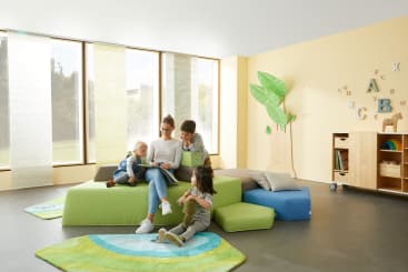 Vier Kinder und ein Erwachsener sitzen zusammen und lesen in einem hellen, modernen Klassenzimmer mit großen Fenstern, hellen Wänden, grünen und blauen Möbeln, einer Baumdekoration an der Wand und Alphabetbuchstaben an der Wand.