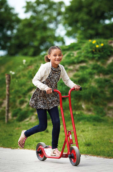 Ein junges Mädchen fährt mit einem roten Motorroller auf einem gepflasterten Weg in einem Park. Sie trägt ein Kleid mit Leopardenmuster, dunkle Leggings, einen hellen Pullover und Sandalen. Im Hintergrund sind grünes Gras und Bäume zu sehen.