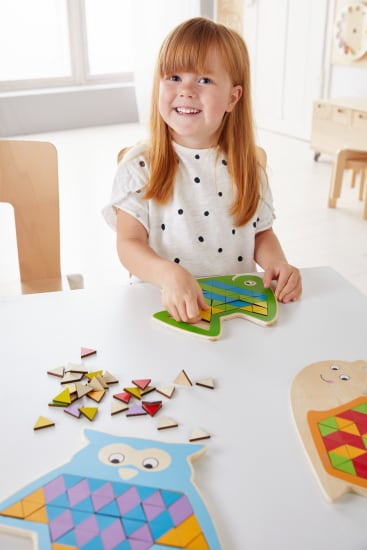 Ein kleines Kind mit roten Haaren und einem weißen gepunkteten Hemd setzt ein hölzernes Eulenpuzzle mit bunten geometrischen Teilen an einem weißen Tisch in einem hellen, kinderfreundlichen Raum zusammen.
