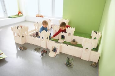 Zwei kleine Kinder spielen mit Spielzeugrittern und -pferden in und um eine große, weiße Spielzeugburg, die auf einem grauen Boden in einem hellen Raum mit grünen Wänden steht.