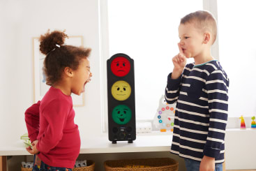 Ein junges Mädchen schreit, während ein Junge in der Nähe steht und einen Finger an seine Lippen hält. Zwischen ihnen steht eine Ampel, die farbige Lichter mit Gesichtern zeigt, in einem hellen Raum mit Kinderspielzeug und Dekorationen.