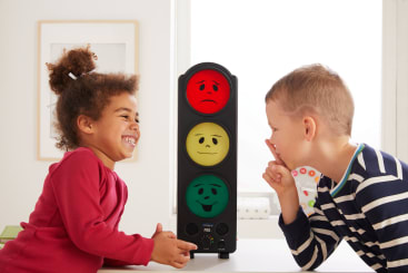 Zwei Kinder sitzen an einem Tisch neben einem elektronischen Gerät mit roten, gelben und grünen Gesichtern. Das Mädchen lächelt, und der Junge macht mit dem Finger an den Lippen eine Geste des Schweigens.