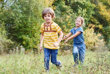 Ein Junge in einem gelben gestreiften Hemd rennt vor ihm her, während ein Mädchen in einem blauen Outfit ihn durch ein grasbewachsenes Feld mit grünen Bäumen im Hintergrund verfolgt. Beide Kinder scheinen im Freien zu spielen.