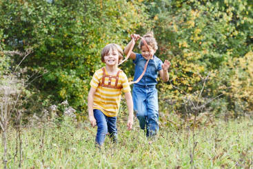 Zwei Kinder laufen durch hohes Gras auf einer Wiese. Das Kind vorne trägt ein gelb gestreiftes Hemd und einen Rucksack, während das Kind hinten, in einem blauen Overall, einen Stock in die Luft hält. Grüne Bäume füllen den Hintergrund.