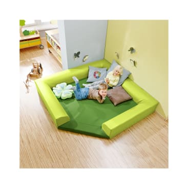 Zwei kleine Kinder liegen auf grünen und gelben gepolsterten Spielmatten in der Ecke eines Raums, der mit Kissen, Wandschmetterlingen und Holzspielzeug auf einem hellen Holzboden dekoriert ist. Im Hintergrund sind Regale mit Behältern zu sehen.
