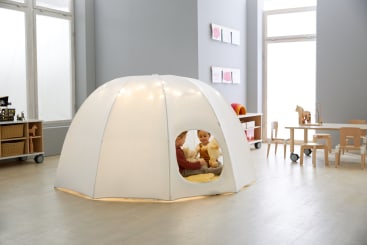 Zwei Kinder sitzen in einem großen, weißen, kuppelförmigen Spielzelt mit Fensteröffnung in einem hellen, modernen Klassenzimmer, das mit Holztischen, Stühlen und Regalen ausgestattet ist.