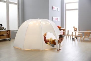 Zwei Kinder spielen in und neben einem großen, weißen, kuppelförmigen Spielzelt in einem hellen, modernen Klassenzimmer mit Holzmöbeln und Spielzeug im Hintergrund.