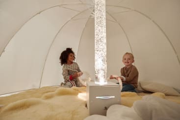 Zwei Kinder sitzen auf einem weichen, gelben Teppich in einem kuppelförmigen Zelt und lächeln sich an. In der Mitte strahlt eine große, beleuchtete Röhre Licht aus. Um sie herum sind Kissen platziert.