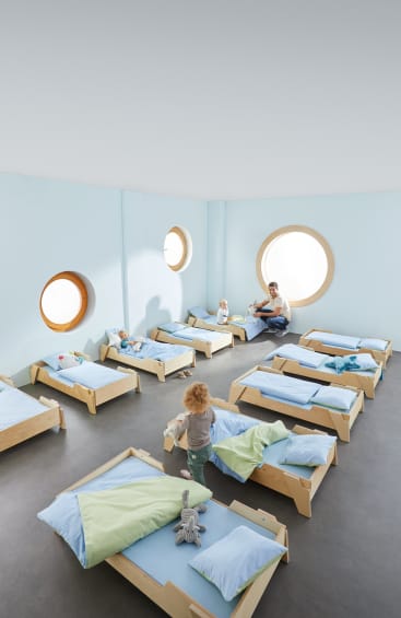 Ein Kinderbetreuungsraum mit hellblauen Wänden, runden Fenstern und zehn kleinen Holzbetten. Es sind mehrere kleine Kinder anwesend; ein Erwachsener sitzt neben einem Kind auf dem Boden. Auf einigen Betten liegen Decken und Spielzeug.