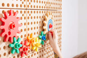 Die Hand eines Kindes dreht ein großes gelbes Zahnrad an einer hellbraunen Wand mit mehreren bunten, ineinander greifenden Zahnrädern in der Nähe. Der Hintergrund ist schlicht und minimalistisch.