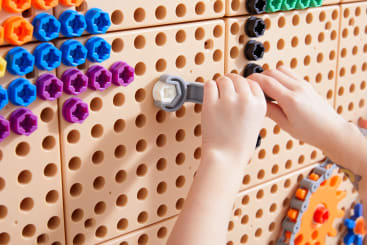 Kinderhände drehen mit einem grauen Spielzeugschlüssel eine weiße Schraube in eine große, hellbraune Stecktafel, die mit bunten sechseckigen Teilen und Zahnrädern gefüllt ist.