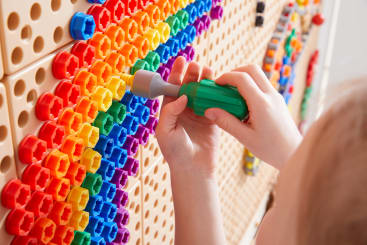 Die Hände eines Kindes setzen mit einem Spielzeugschraubendreher farbige Kunststoffbolzen in eine vertikale, in Reihen von Regenbogenfarben angeordnete Stecktafel ein. Nur die Hände und ein Teil des Schraubenziehers sind sichtbar.