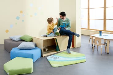 Ein Erwachsener und ein Kind sitzen zusammen in einem hell erleuchteten Spielzimmer, das mit bunten Kissen, einem Teppich und kleinen Möbeln ausgestattet ist. Der Erwachsene liest dem Kind ein Buch vor, während beide auf einem Holzgestell an der Wand sitzen.