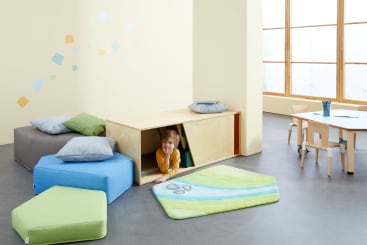 Ein Kind lächelt, während es in einem hellen Raum mit großen Fenstern, einer verzierten Wand und einem kleinen Tisch mit Stühlen aus einem hölzernen Spielgerät krabbelt, das von bunten Kissen umgeben ist.