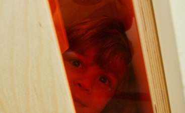 Das Gesicht eines Kindes ist teilweise durch eine rot gefärbte transparente Fläche sichtbar, die von hellen Holzplatten umgeben ist. Das Kind blickt mit einem neutralen Ausdruck nach außen.
