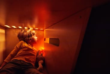 Ein kleines Kind in einem gelben Hemd sitzt in einem kleinen hölzernen Raum, der durch Löcher und einen Schlitz von warmem orangefarbenem Licht erhellt wird, und hält ein Stofftier in der Hand, das in Richtung des Lichts schaut.