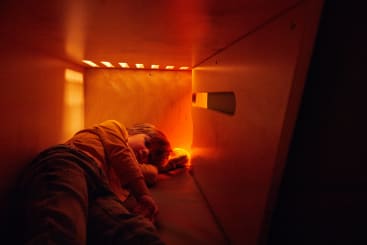 Ein Kind liegt in einem kleinen, hölzernen, kastenähnlichen Raum mit warmer, orangefarbener Beleuchtung, hält ein leuchtend orangefarbenes Objekt in der Hand und schaut nach vorne.
