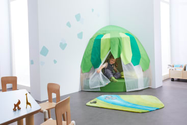 Ein Kind sitzt in einem Spielzelt aus grünem und weißem Stoff, das in der Ecke eines hellen Raums steht. In der Nähe befinden sich ein bunter Teppich, Holzstühle, ein Tisch mit Tierspielzeug und ein Rollwagen mit Spielzeug im Hintergrund.