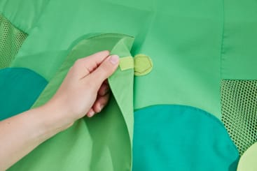 Eine Hand zieht eine grüne Stofflasche mit einem Klettverschluss zurück, so dass darunter ein hellerer grüner Stoff zum Vorschein kommt. Der Hintergrund besteht aus verschiedenen Grüntönen von Stoff und Netz.