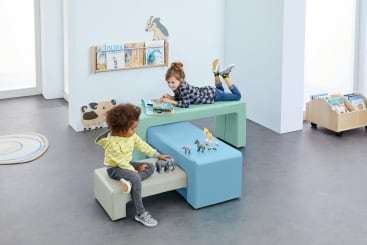 Zwei Kinder spielen in einem hellen Raum; eines liest ein Buch auf einer pastellgrünen Bank, während das andere auf einem beigen Hocker sitzt und mit Tierfiguren auf einem blauen Tisch spielt. Im Hintergrund sind Bücherregale und Spielzeug zu sehen.