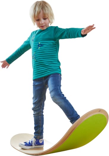 Ein Kind mit blondem Haar, das ein grün gestreiftes Hemd, Jeans und blaue Turnschuhe trägt, balanciert mit ausgestreckten Armen auf einem geschwungenen Balancebrett aus Holz mit grüner Oberfläche.