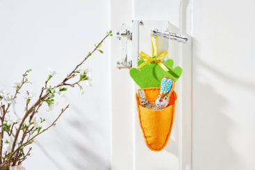 An einer weißen Türklinke hängt ein Filzbeutel in Form einer Karotte, gefüllt mit Osterpralinen, darunter ein Hase und ein Küken; links an einer weißen Wand ist ein blühender Zweig zu sehen.