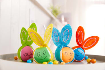Vier bunte Eier mit aufgemalten Gesichtern und Hasenohren aus Filz liegen auf einem Tisch, umgeben von kleinen bunten Süßigkeiten, vor einem hellen, verschwommenen Hintergrund.