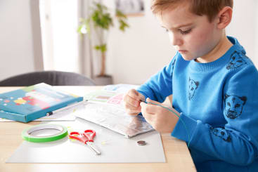 Ein kleines Kind in einem blauen Sweatshirt sitzt an einem Tisch und arbeitet an einem Bastelprojekt mit Alufolie, Schere, Klebeband, einer Knopfzellenbatterie und einem grünen Draht. Auf dem Tisch sind verschiedene Materialien und Anleitungen zu sehen.