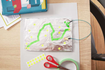 Ein Stück Folie mit grünem Klebeband und gelben Punktaufklebern bildet einen einfachen Stromkreis auf einem Schreibtisch, umgeben von einer Schachtel, Papier, grünem Klebeband, einer roten Schere und einem Stuhl.