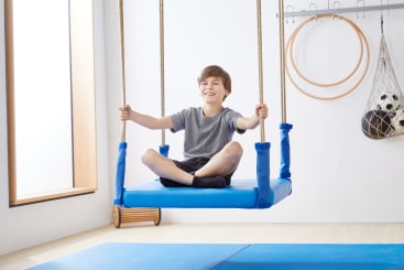 Ein Junge in einem grauen T-Shirt und Shorts sitzt im Schneidersitz auf einer blauen Plattformschaukel in einer Halle und hält sich mit beiden Händen an den Seilen fest. Im Hintergrund sind Gymnastikgeräte und ein Netz mit Bällen zu sehen.