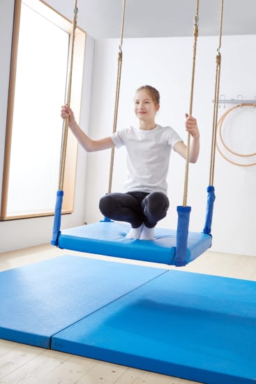 Ein Kind, das ein weißes T-Shirt und eine dunkle Hose trägt, sitzt im Schneidersitz auf einer aufgehängten Schaukel mit blauen Polstern über blauen Turnmatten in einem hellen Innenraum.
